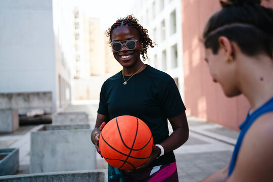 A smiling African American woman with braided hair and sunglasses holds a basketball. She is outdoors in an urban setting, looking at the camera, ready to play. Another person is partially visible.