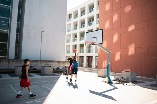 Three young adults play basketball on an outdoor court. Two players jump for the ball near the hoop, while another watches. Modern buildings frame the urban scene.