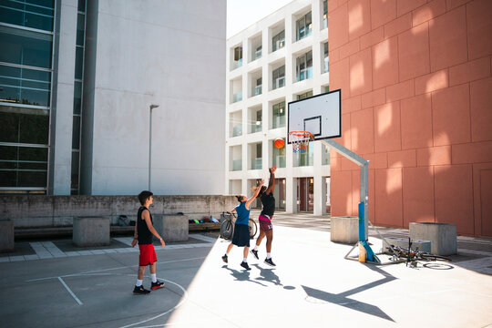 Three young people play basketball on an outdoor court. Two players jump for the ball near the hoop, one shooting and one defending. A third person observes the game.