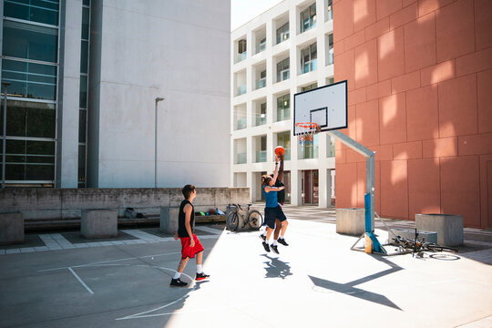 Two young men play an energetic game of basketball on an outdoor court. One player leaps high to shoot the ball towards the hoop as the other watches intently. Urban buildings surround the court.