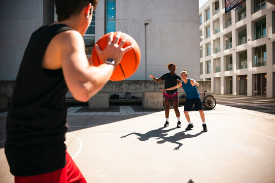 Three young adults play basketball on an outdoor urban court. One player holds the ball, preparing to shoot or pass, while two opponents actively defend. They enjoy sports on a sunny day.