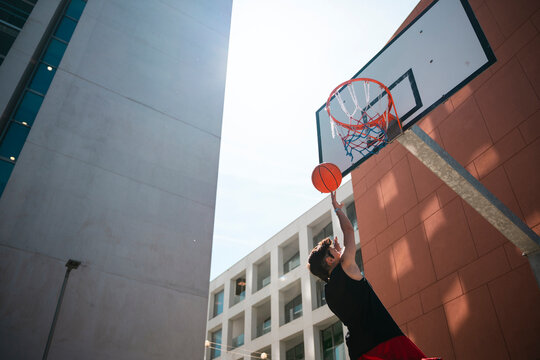 A young man plays basketball outdoors under bright sunlight. He reaches high to dunk the ball into the hoop, demonstrating athletic skill and urban sports activity.