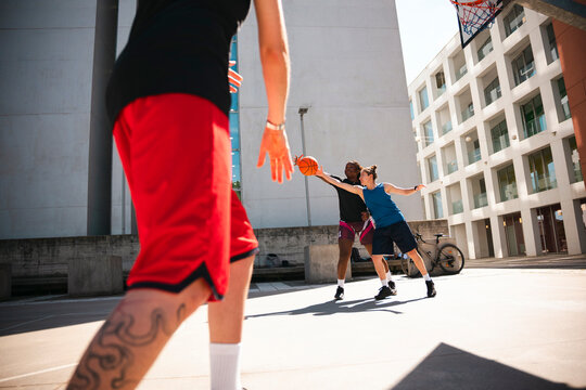 Three diverse friends play an intense game of basketball on an outdoor court. Two players reach for the ball while another approaches the action.