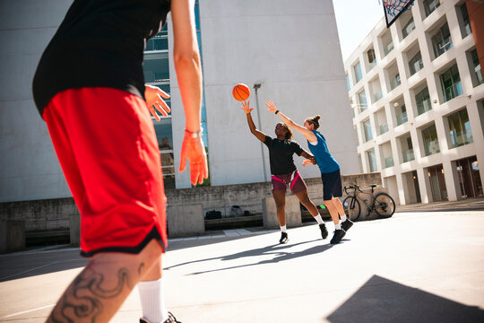 Three people play basketball on an outdoor court. A player attempts a shot while a defender blocks, with another player in the foreground. They enjoy the game on a sunny day.