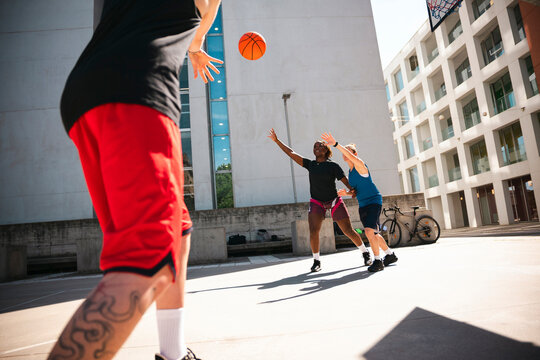 Three diverse players enjoy an energetic outdoor basketball game. One person passes the ball as two opponents reach to intercept it on a sunny urban court.