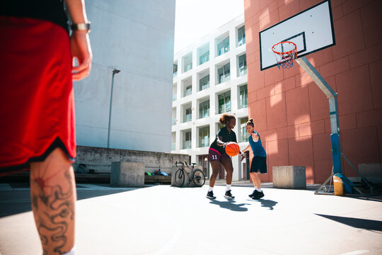 Two women play basketball on an outdoor court under a clear sky. One woman dribbles the ball, while the other defends. A third person observes from the foreground.