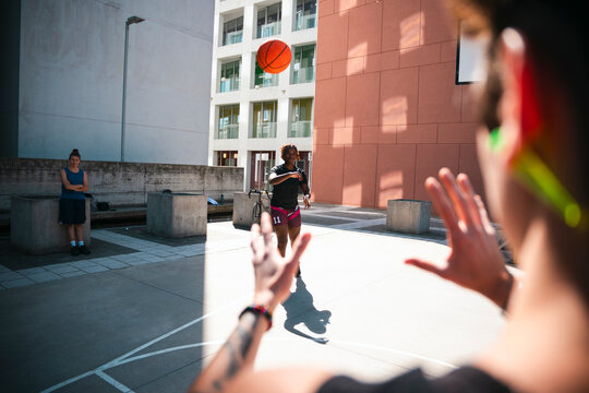 Three individuals engage in an outdoor basketball game. A player in the foreground reaches for the ball thrown by another, while a third person observes the action on the urban court.