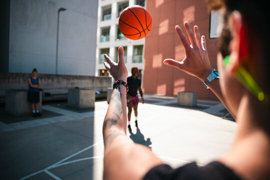 A person with a tattooed arm and a watch reaches for a basketball in mid-air on an urban outdoor court. Other players are present in the background. The sun shines brightly.