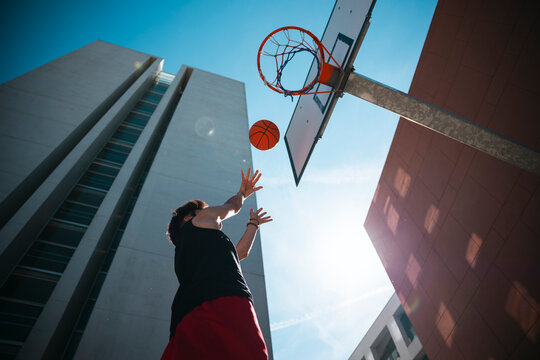 A person in athletic wear shoots a basketball towards the hoop. Tall, modern buildings frame the outdoor court under a bright, sunny sky. A strong lens flare is visible.