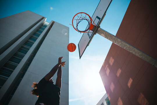A silhouetted person shoots a basketball towards the hoop on an outdoor court. Modern buildings frame the bright blue sky, with the sun creating a lens flare.