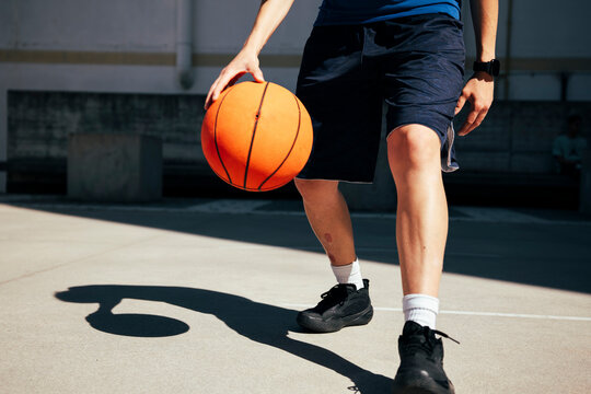 A young person dribbles an orange basketball on an outdoor court. The player wears athletic shorts and sneakers, practicing their skills under the bright sun.