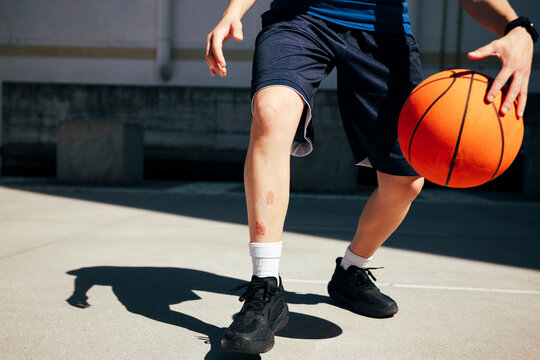 A basketball player moves across an outdoor court, dribbling the ball. A scrape on their leg highlights the physical nature of the sport and their commitment.