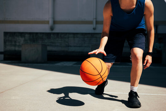 A person in athletic wear dribbles an orange basketball on a sunny outdoor court. The player's hand is above the ball, mid-dribble, showcasing active play.