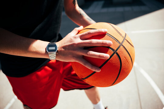 A player holds a basketball on an outdoor court, hands gripping the textured surface. A digital watch is on their wrist. This image conveys readiness and passion for the game.