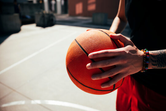 A young athlete holds a textured basketball firmly with both hands on an outdoor court. The person wears a black shirt and red shorts, showcasing a casual, active lifestyle.