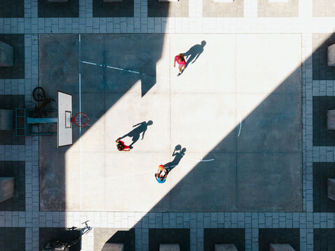 Three people play basketball on an outdoor court under bright sunlight, casting long shadows. One person dribbles, another holds the ball, and a third stands nearby.