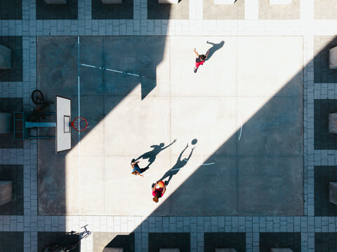 Three young adults play basketball on an outdoor court. One person dribbles the ball while another reaches for it. A third person runs across the court. Sunlight casts long shadows.