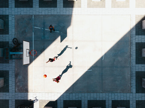 Three young people play basketball on an outdoor court. Two actively engage with the ball, one dribbling and another reaching. A third person watches from the side. Sunlight casts long shadows.