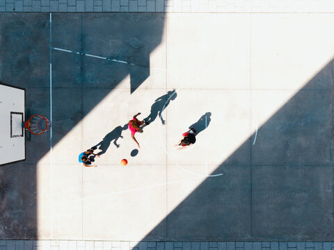 Three young people play basketball on an outdoor court. One dribbles the ball, another defends, and a third player watches. Strong sunlight casts long shadows across the concrete surface.