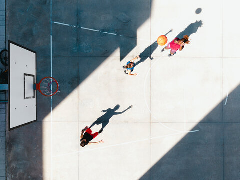Three young adults play basketball on an outdoor court under bright sunlight. One person dribbles the ball while two others move around the court. Long shadows stretch across the concrete.