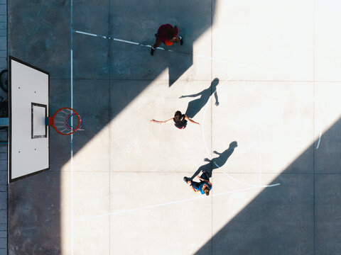 Three young adults play basketball on an outdoor court under bright sunlight. One player holds the ball near the hoop, while others move around. Strong shadows stretch across the concrete.