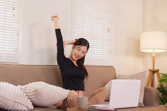 Asian woman stretching arms working from home office with laptop computer on sofa feeling happy relaxed during break time remote work freelance lifestyle