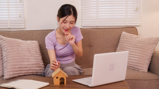 Asian women planning home purchase using laptop computer for real estate research sitting on sofa with miniature house model at modern living room interior