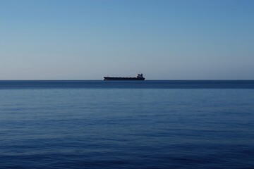 Obraz premium Large cargo ship sails on calm blue ocean under clear sky. The vessel travels on horizon line. Marine shipping and international trade themes.