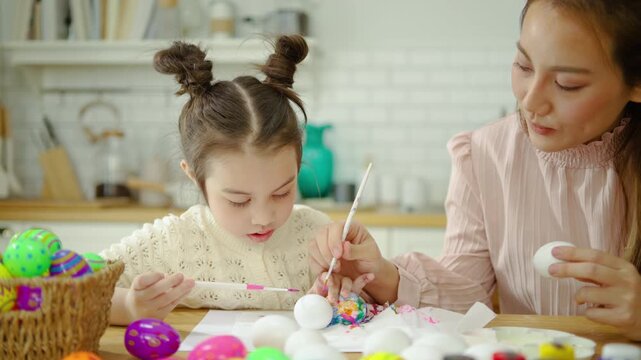 Loving Asian mother and her cute young daughter enjoy decorating colorful Easter eggs together at a bright kitchen table, celebrating spring holiday with a fun family creative art activity.