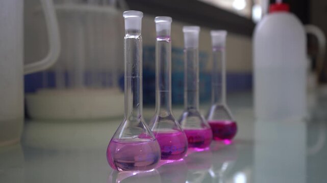 Close-up shot of four volumetric flasks containing a bright pink chemical solution lined up on a reflective lab bench. Ideal B-roll for scientific research, chemistry, and pharmaceutical development.