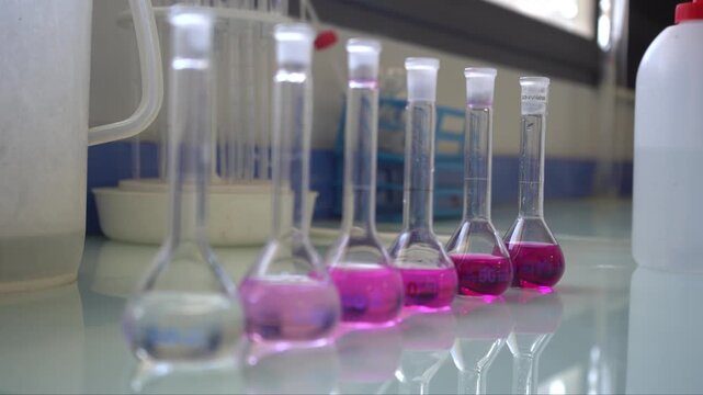 A scientist places a volumetric flask into a row showing an acid-base titration color gradient. Ranging from clear to deep pink, this B-roll perfectly illustrates water alkalinity testing and chemistr