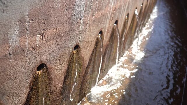 Close-up of clarified water trickling through concrete weir holes into a sunlit trough at a treatment plant. Features authentic audio. Ideal B-roll for water purification and infrastructure projects.
