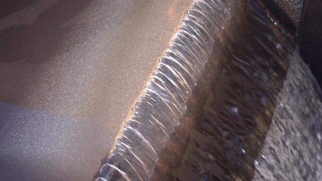 Extreme close-up of a smooth, glass-like sheet of water flowing over a weir edge into a turbulent basin. Features authentic rushing audio. Ideal B-roll for fluid dynamics and water treatment projects.