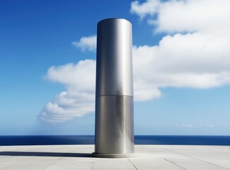 Obraz premium Tall metal cylinder stands on paved area near ocean under blue sky with white clouds. Modern architecture piece against sea horizon. Minimalist design element.