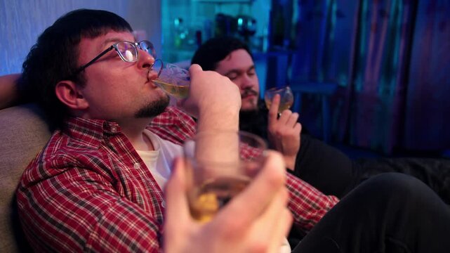 Two relaxed fiends relax with drinks and friendly talk. Young caucasian men sit in living room, holds glass with alcohol beverages and engaging in friendly conversation. The company is relaxing