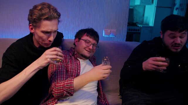 Casual evening with young funny drunk friends on sofa. Relaxed evening where caucasian men enjoy alcohol beverages. Comfortable company in dimly cozy room. The harm of alcohol consumption