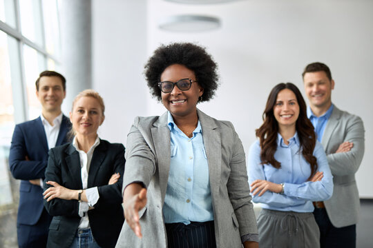 Happy black female leader offering a handshake in the office. Her colleagues are in the background