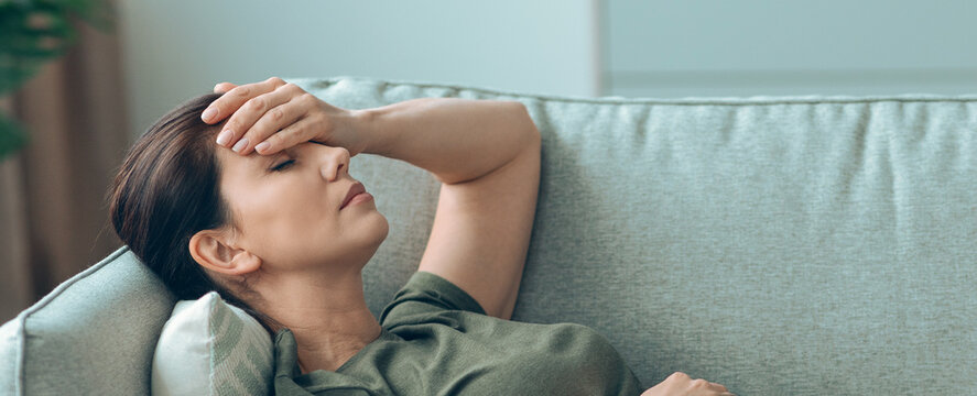 Woman lies down on couch with her eyes closed, touching her forehead with one hand. She appears to feel unwell in a simple living space with natural light.