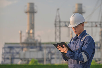 Fototapeta premium Asian male electrical engineer holding digital tablet standing at power plant station. Professional energy industry technician working on infrastructure maintenance and renewable resource project.