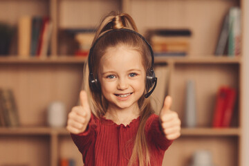 A girl with long hair smiles while giving two thumbs up. She wears a headset and stands in a room with shelves filled with books. The atmosphere suggests she is engaged and happy. © Prostock-studio