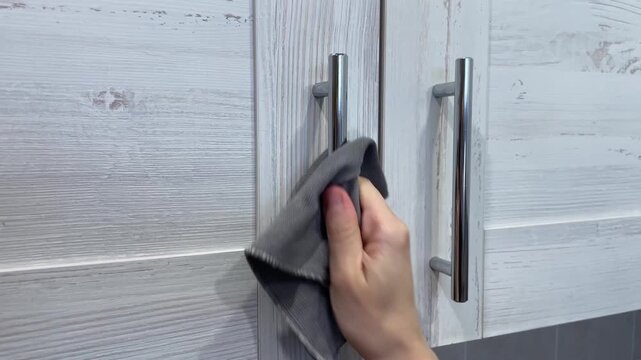 Closeup hands cleaning cabinet handles grey wood panels and brushed metal pulls wiped with cloth to remove fingerprints and smudges, kitchen refresh and hardware care, neat modern styling