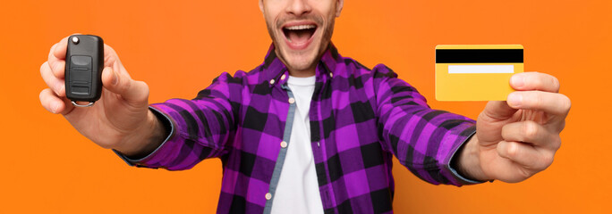 A man is smiling widely while holding a car key in one hand and a credit card in the other. He stands in front of an orange wall, showing clear excitement about his purchase or ownership. © Prostock-studio