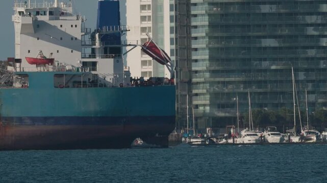 Cargo ship docks at the harbor near city buildings and yachts in bright weather