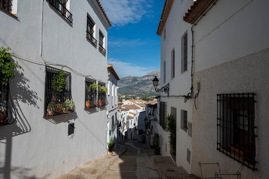 Narrow street with white houses in the old town of Altea, Alicante, Spain, with Mediterranean architecture and mountain landscape in the background on a sunny day.