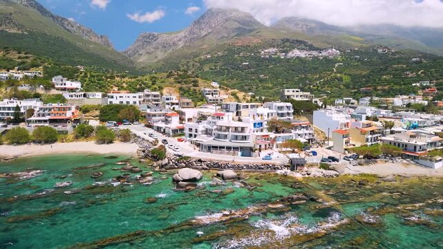 Plakias South Crete Greece drone aerial low flight from sea reveal coastal town with white houses and turquoise transparent water, mountain background stones in sea wide angle panorama