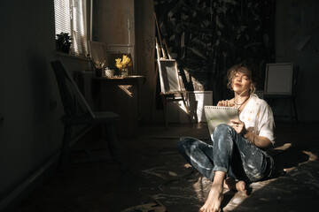 Young curly woman artist drawing on notebook sitting on floor at art studio in Hard light, sunny © Andrii Lysenko