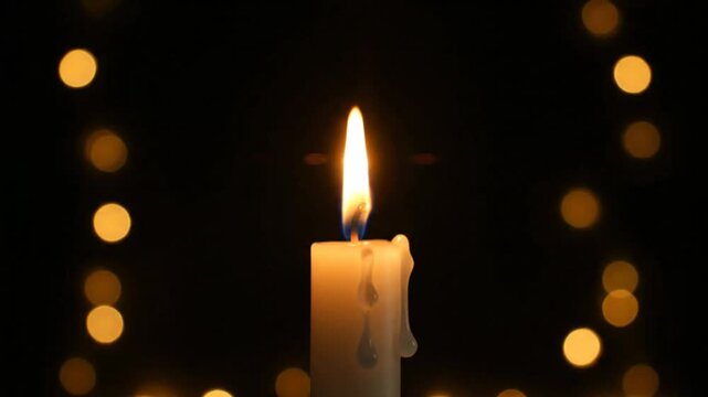 Burning candle with a small flame against a dark background with festive bokeh lights creating a peaceful, warm atmosphere.