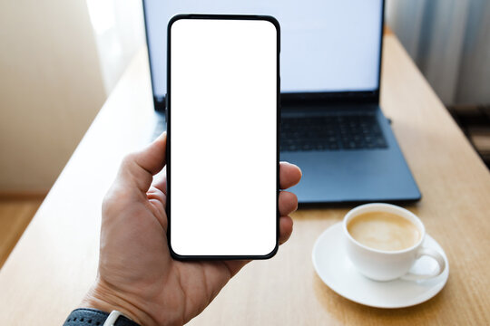  Closeup Hand Holding Smartphone with Blank White Screen Against Blurred Background with Laptop and Cup of Coffee on the Table