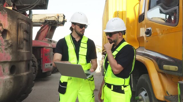 Two engineers in safety vests analyze laptop data near cargo truck and container, coordinating logistics and industrial transport operations.