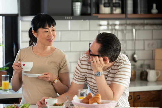 Asian woman enjoying coffee while talking with her partner during breakfast in a modern kitchen. Relaxed morning routine, cozy home atmosphere, connection and everyday lifestyle